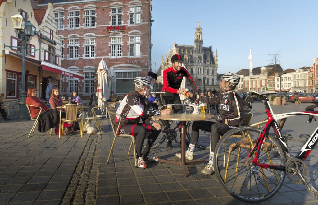Drei Radfahrer sitzen in einem Café auf dem Stadtplatz und stoßen mit Bier an, lächelnd, mit ihren Fahrrädern in der Nähe.